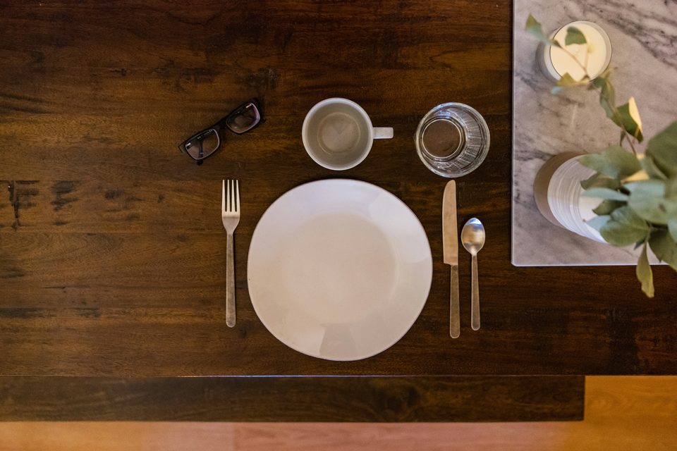 Mesa de madera oscura puesta con un plato blanco, cubiertos, una taza y un vaso, vista desde arriba.
