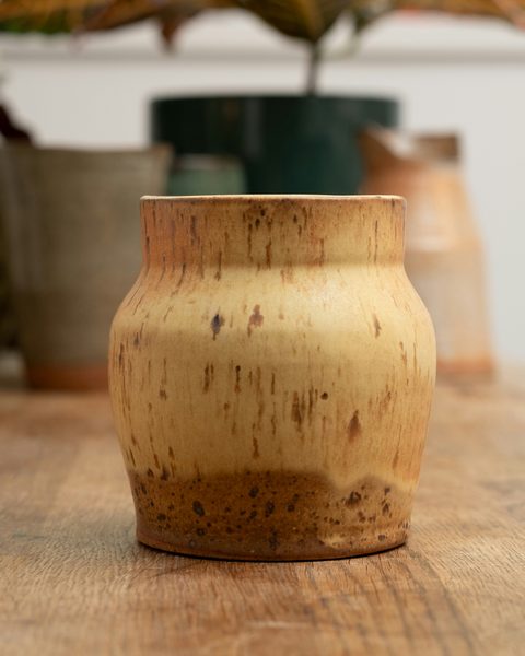 Maceta de cerámica color miel con motas y el pie de barro sin esmaltar, sobre una mesa de madera.