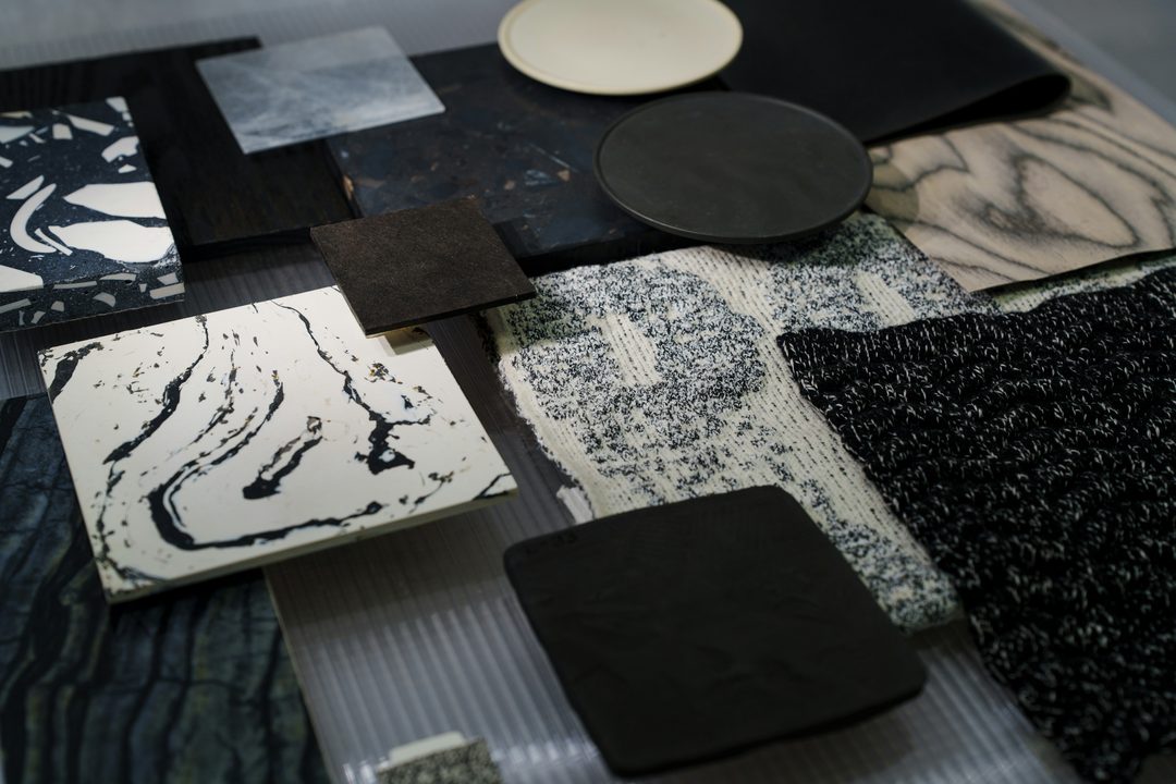 Muestras de materiales sobre una mesa: terrazos, piedras, madera, cerámica y textiles en blanco y negro.