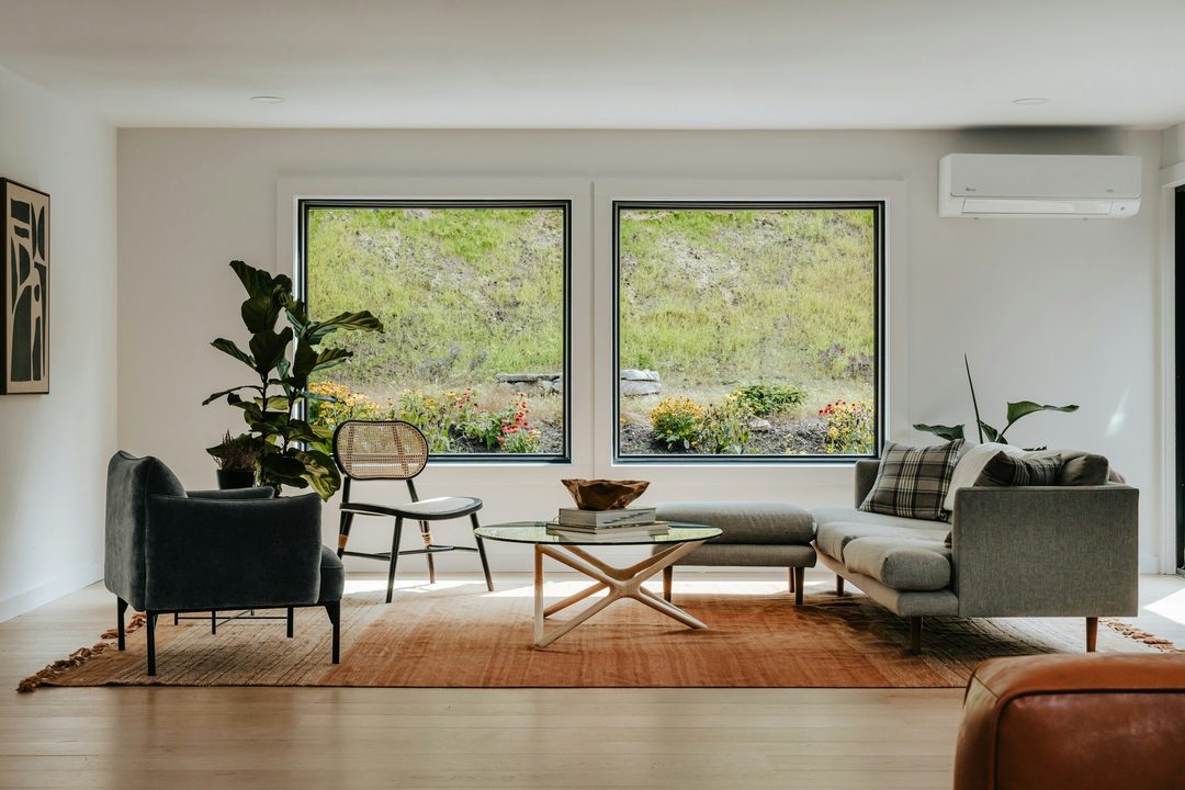 Sala luminosa con dos ventanas cuadradas hacia un talud con flores, un sofá gris, sillones y una mesa de centro sobre un tapete terracota.