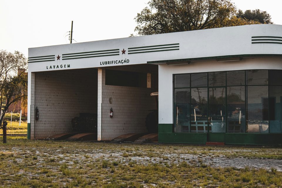 Fachada blanca y verde de un antiguo taller de servicio, con dos bahías abiertas, rótulos pintados de lavado y lubricación y un ventanal.