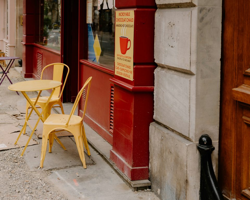 Mesa y dos sillas amarillas sobre la banqueta, frente al aparador rojo de una cafetería.